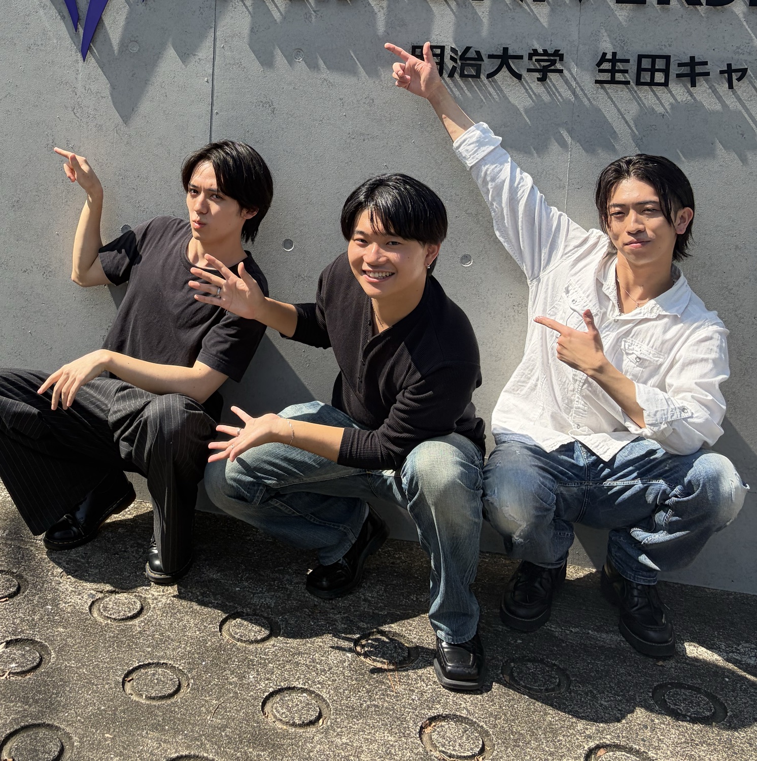 明治大学の壁の前でポーズをとるSARACHIのメンバー3人
